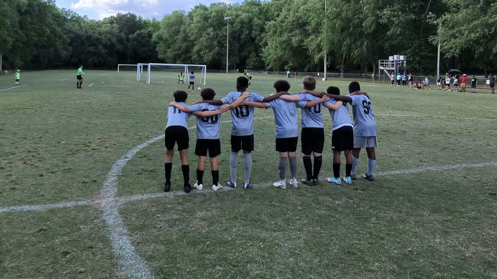 Seven youth players in light blue kit standing arm in arm in a huddle on the pitch, backs to the camera