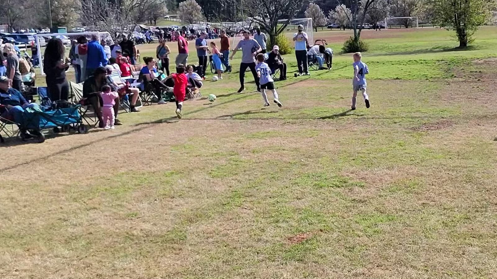 Families set up in camp chairs along the sideline of a youth soccer field, with a game in progress and cars parked behind
